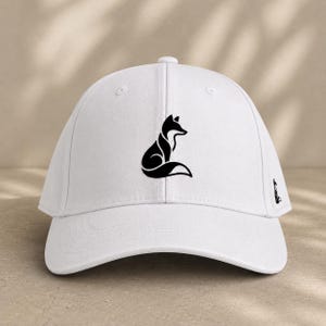 Pode incluir: Bon&eacute; de beisebol branco com uma silhueta de raposa preta na frente e um log&oacute;tipo de raposa menor na lateral. O bon&eacute; tem uma aba curva e uma al&ccedil;a ajust&aacute;vel. Um acess&oacute;rio elegante para atividades ao ar livre.