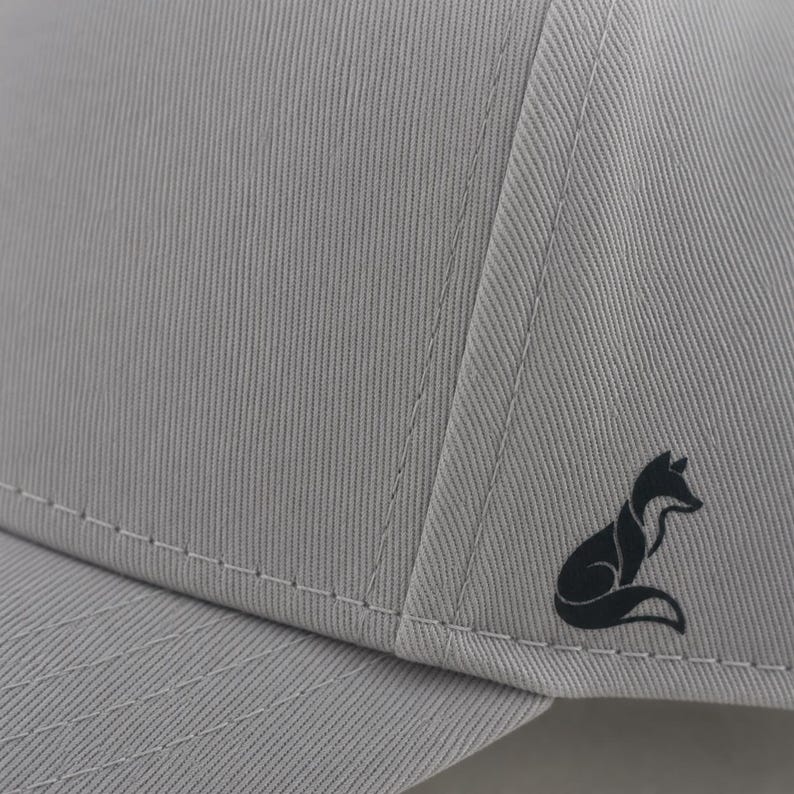 Pode incluir: Bon&eacute; de beisebol cinza claro com um logotipo de raposa preta na lateral. O bon&eacute; tem um design estruturado com costuras vis&iacute;veis e uma aba curva. O tecido tem uma textura vertical sutil.