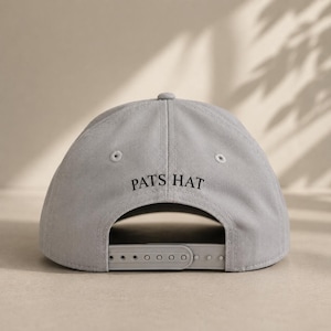 Pode incluir: Bon&eacute; de beisebol cinza claro com o texto "PATS HAT" em preto. O bon&eacute; tem uma aba curva e uma al&ccedil;a ajust&aacute;vel na parte de tr&aacute;s. Feito de um material macio e dur&aacute;vel.