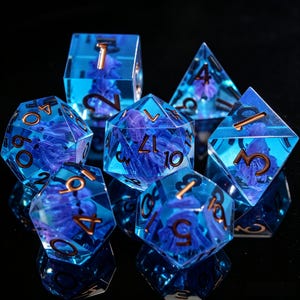 Puede incluir: Un juego de siete dados poliédricos hechos de resina azul translúcida, cada uno con un elemento floral púrpura suspendido. Los dados presentan números grabados rellenos con un pigmento de color cobre metálico, visibles en todas las caras.