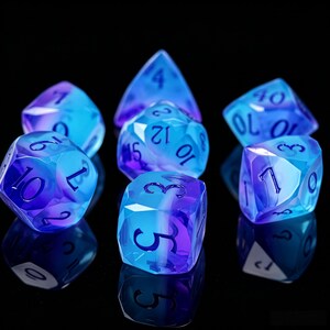 Peut inclure: Un ensemble de sept dés polyédriques présentant un dégradé translucide de couleurs bleu et violet. Chaque dé possède des bords tranchants et des chiffres foncés gravés sur les faces. La collection comprend diverses formes pour les jeux.