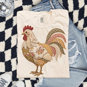 Könnte beinhalten: Cremefarbenes T-Shirt mit einem bunten Hahn-Design. Der Hahn besteht aus verschiedenen gemusterten Stoffen, darunter Blumen-, Tupfen- und Streifenmuster. Der Hahn hat einen roten Kamm und Kehllappen. Das T-Shirt ist auf einer Jeans gefaltet.