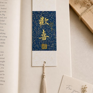 May include: A rectangular paper bookmark featuring a central dark blue panel with gold speckled details and gold Chinese calligraphy characters. The bookmark has cream-colored ends, a small hole at the bottom, and a beige silk tassel attached.