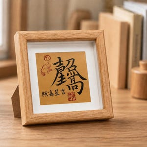 The tabletop calligraphy frame standing on a wooden desk beside a small potted plant, with blurred books in the background, showing how it looks displayed in a home office or study.