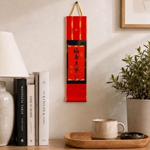 Mini red Chinese calligraphy wall scroll displayed in a cozy home setting with books, lamp, plant, and wooden shelf decor