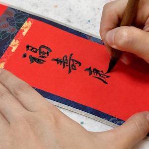 Close-up process image of hand-written Chinese calligraphy on red paper for a personalized mini wall scroll