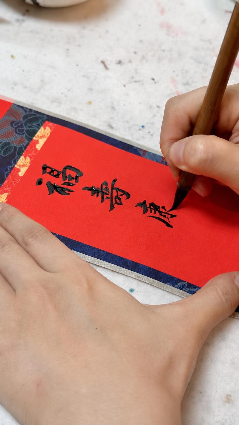 Close-up process image of hand-written Chinese calligraphy on red paper for a personalized mini wall scroll