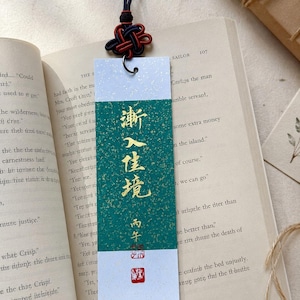 May include: A rectangular paper bookmark featuring a central green panel with gold flecks and gold Chinese calligraphy characters. The bookmark has white top and bottom borders, a metal eyelet, and a decorative black and red knotted cord.