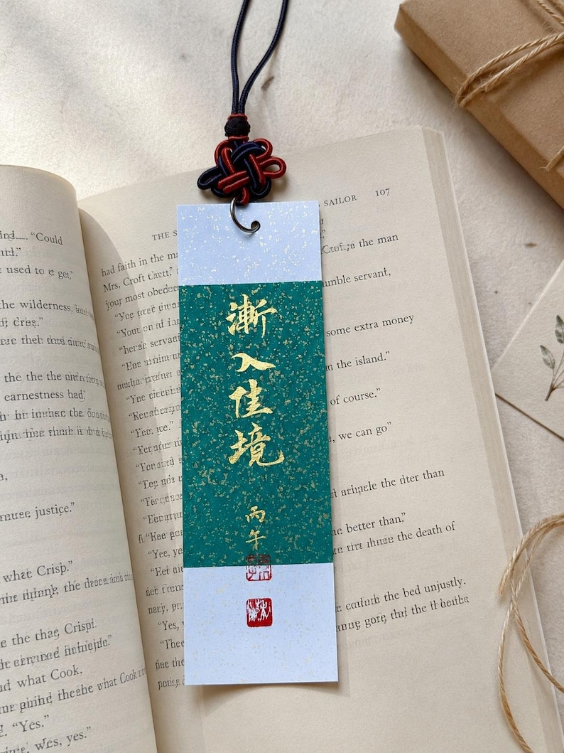 May include: A rectangular paper bookmark featuring a central green panel with gold flecks and gold Chinese calligraphy characters. The bookmark has white top and bottom borders, a metal eyelet, and a decorative black and red knotted cord.