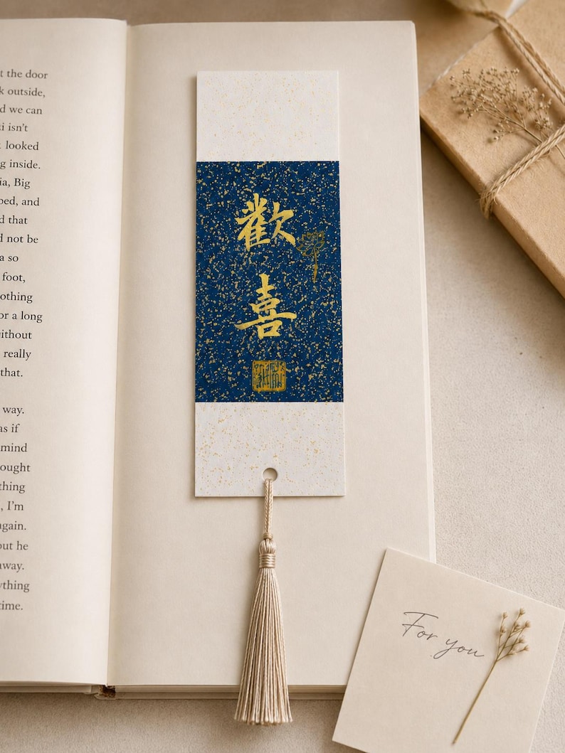 May include: A rectangular paper bookmark featuring a central dark blue panel with gold speckled details and gold Chinese calligraphy characters. The bookmark has cream-colored ends, a small hole at the bottom, and a beige silk tassel attached.