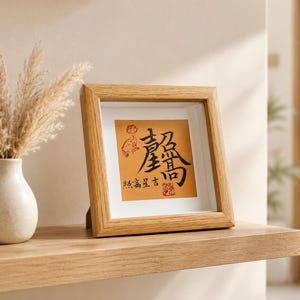 The tabletop calligraphy frame displayed on a floating wood shelf near an entryway, next to a ceramic vase with dried pampas grass, in soft natural daylight.