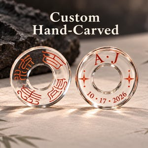Op de afbeelding: Twee ronde, transparante glazen ringen met aangepaste rode gravures. De ene ring toont vier verschillende Aziatische karakters, terwijl de andere de initialen A.J, twee vierpuntige sterren en de datum 10.17.2026 in een strak lettertype bevat.