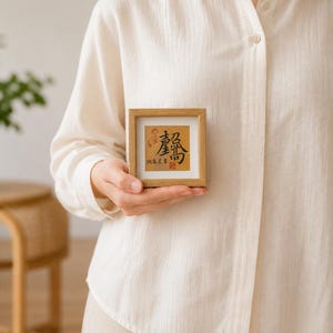 A hand holding the tabletop calligraphy frame at chest height against a soft neutral background, showing its true size relative to an adult hand.