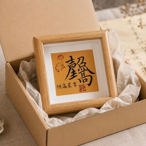 The tabletop calligraphy frame nestled in an open kraft gift box lined with tissue paper, shown as it would arrive when gifted, with calligraphy brushes visible nearby.