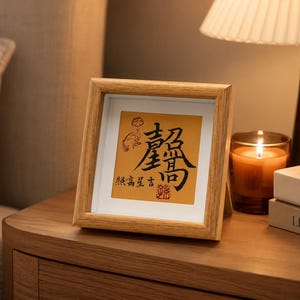 The tabletop calligraphy frame standing on a bedside table next to a lit candle, a warm lamp, and a small stack of books, in soft evening light.
