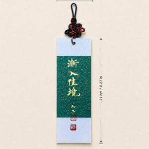 May include: A rectangular bookmark featuring a central green panel with gold flecks and gold Chinese calligraphy, bordered by white sections. It includes a dark blue and red decorative knot at the top. Dimensions are 7 cm wide by 21 cm long.