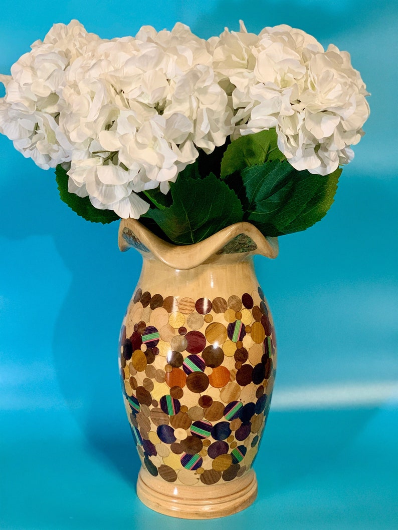 Puede incluir: Un ramo de hortensias blancas en un jarr&oacute;n de madera con un dise&ntilde;o de mosaico &uacute;nico. El jarr&oacute;n est&aacute; hecho de diferentes piezas de madera en tonos marrones, morados y verdes, creando un patr&oacute;n circular.