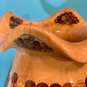 Puede incluir: Un jarr&oacute;n de madera con un dise&ntilde;o &uacute;nico y ondulado. El jarr&oacute;n est&aacute; hecho de madera clara con detalles marrones oscuros y un patr&oacute;n circular alrededor de la base.