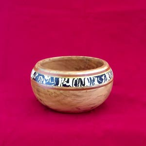 May include: A wooden bowl with a black, white, and gold inlay design around the rim. The bowl is made of light brown wood and has a natural finish.