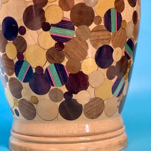Puede incluir: Un jarr&oacute;n de madera con un dise&ntilde;o &uacute;nico. El jarr&oacute;n est&aacute; cubierto de un mosaico de peque&ntilde;as piezas de madera redondas en varios tonos de marr&oacute;n, beige y morado. El jarr&oacute;n tiene una base lisa y redondeada.