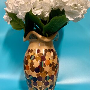 Puede incluir: Un jarr&oacute;n de madera con un dise&ntilde;o &uacute;nico que presenta un mosaico de peque&ntilde;as piezas de madera redondas en varios tonos de marr&oacute;n, naranja y morado. El jarr&oacute;n est&aacute; lleno de flores de hortensia blancas.