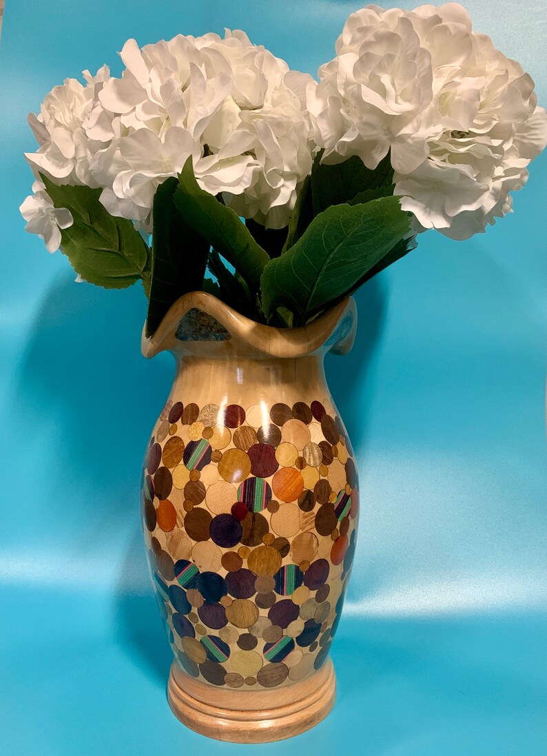 Puede incluir: Un jarr&oacute;n de madera con un dise&ntilde;o &uacute;nico que presenta un mosaico de peque&ntilde;as piezas de madera redondas en varios tonos de marr&oacute;n, naranja y morado. El jarr&oacute;n est&aacute; lleno de flores de hortensia blancas.