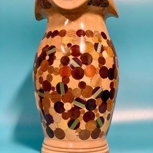Puede incluir: Un jarr&oacute;n de madera con un dise&ntilde;o &uacute;nico. El jarr&oacute;n est&aacute; hecho de madera y tiene una parte superior ondulada. El cuerpo del jarr&oacute;n est&aacute; cubierto de un mosaico de peque&ntilde;as piezas de madera redondas en varios tonos de marr&oacute;n, beige y negro. El jarr&oacute;n tiene una base marr&oacute;n claro.