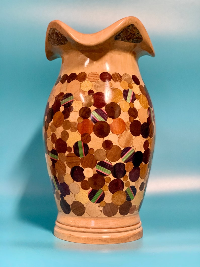 Puede incluir: Un jarr&oacute;n de madera con un dise&ntilde;o &uacute;nico. El jarr&oacute;n est&aacute; hecho de madera y tiene una parte superior ondulada. El cuerpo del jarr&oacute;n est&aacute; cubierto de un mosaico de peque&ntilde;as piezas de madera redondas en varios tonos de marr&oacute;n, beige y negro. El jarr&oacute;n tiene una base marr&oacute;n claro.