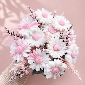 Peut inclure: Un bouquet de fleurs en forme de marguerites blanches et roses, avec des centres roses et des tiges délicates. Les fleurs sont disposées en bouquet, enveloppées dans du papier rose, sur fond rose.
