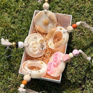 May include: Four circular wax air fresheners with dried flowers and beads in a white box. Each piece features a hanging loop with wooden and colored beads. One wax piece is pink with floral details, while others are cream with botanical accents.