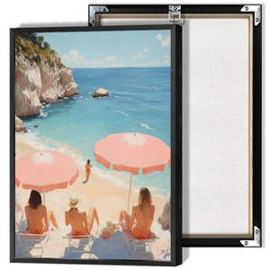 May include: A framed canvas print depicts a sunny beach scene. Three figures sit in beach chairs under a pink umbrella, facing the ocean. The artwork features a turquoise sea, a sandy beach, and a rocky cliffside. The canvas is framed in black.