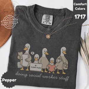 May include: A charcoal gray t-shirt featuring a graphic of five cartoon ducks engaged in office tasks. One duck holds a clipboard, another uses a laptop labeled "CLIENT FILES," and one holds a heart. The text "doing social worker stuff" is printed below.
