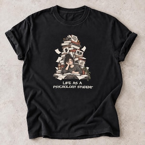May include: A black short-sleeved t-shirt featuring a graphic of a person sitting at a desk surrounded by a large stack of books. The text LIFE AS A PSYCHOLOGY STUDENT is printed in white below the illustration, which includes an hourglass.