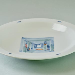May include: A white ceramic bowl featuring a central square design with blue, teal, and orange watercolour-style brushstrokes in a spiral pattern. The rim of the bowl is decorated with small, evenly spaced blue diamond-shaped accents.