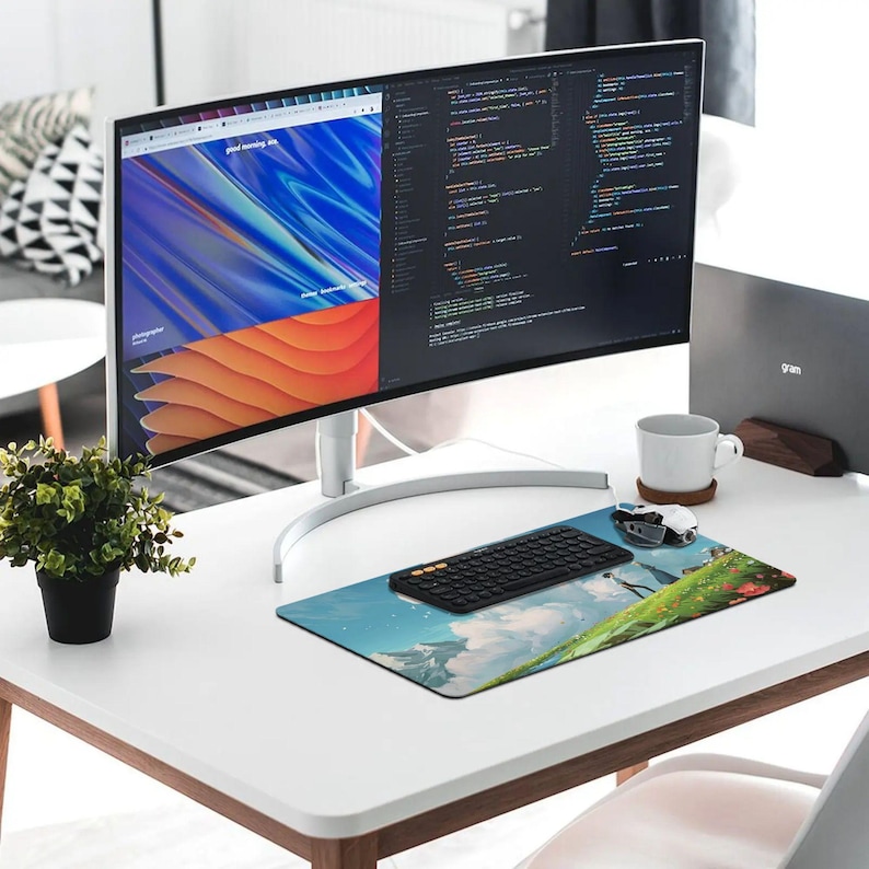 K&ouml;nnte beinhalten: Ein moderner Arbeitsplatz mit einem gebogenen Computermonitor, der Website-Design und Code anzeigt. Eine schwarze Tastatur, Maus und ein Mauspad mit Landschaftsdesign befinden sich auf dem wei&szlig;en Schreibtisch. Eine kleine Topfpflanze und eine wei&szlig;e Tasse vervollst&auml;ndigen das Setup.