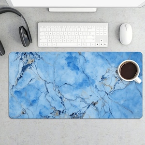 May include: A rectangular desk mat with a blue marble design. The mat has a white keyboard, mouse, and a cup of coffee on it. A pair of black headphones is on the left side of the mat.