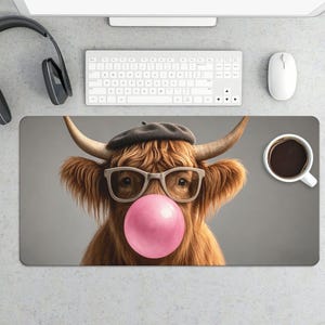 May include: A rectangular mousepad featuring a Highland cow wearing glasses, a beret, and blowing a pink bubble. A white coffee mug with black coffee sits to the right. A keyboard, mouse, and headphones are visible.