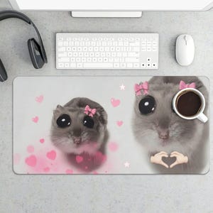 May include: A rectangular mousepad featuring a cute hamster design with large eyes and a pink bow. The design includes pink hearts and a cup of coffee. A keyboard, mouse, and headphones are visible in the background.