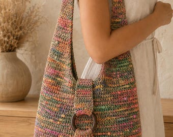 May include: A hobo-style shoulder bag crafted from multicolored variegated yarn in shades of pink, green, orange, and blue. The bag features a single wide strap, a central flap closure secured with a dark brown wooden ring, and a matching yarn tassel.