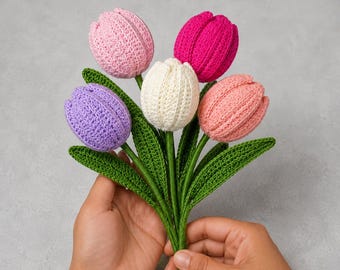 May include: A bouquet of five crocheted tulips featuring yarn petals in shades of light pink, magenta, white, lavender, and peach. Each flower is attached to a green crocheted stem with long, pointed green leaves, all crafted with a visible yarn texture.