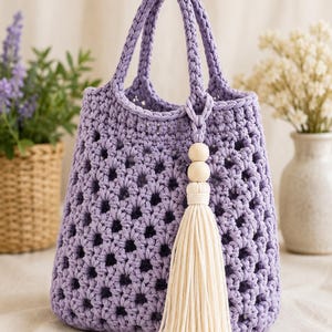 May include: A lavender crochet handbag featuring an openwork diamond pattern, two braided top handles, and a decorative accessory consisting of two round wooden beads attached to a long, cream-colored cotton cord tassel hanging from one handle.