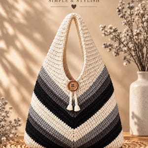 Peut inclure: Un sac à main en crochet de forme triangulaire avec un motif chevron composé de rayures alternées crème, gris clair, gris foncé et noir. Le sac possède une anse intégrée, un bouton en bois central et deux petits pompons couleur crème.