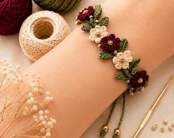 May include: A crochet bracelet featuring alternating dark red and cream-coloured flowers with green leaves and small gold beads. The bracelet has two dangling green cords with gold bead ends, and is displayed alongside yarn balls and a gold crochet hook.