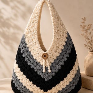 May include: A crochet hobo bag featuring a chevron pattern in cream, grey, and black yarn. The bag has a single integrated handle and a central wooden button closure with two dangling cream-colored cords, creating a textured, triangular silhouette.