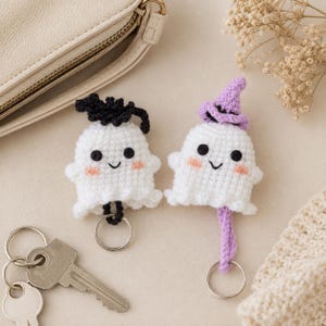 May include: Two crocheted ghost keychains made of white yarn with black embroidered eyes, small smiles, and pink cheeks. One ghost wears a black crocheted hat, and the other wears a lavender crocheted hat. Each ghost is attached to a silver metal keyring.