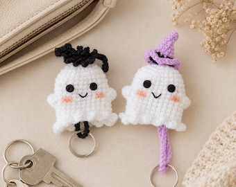 May include: Two crocheted ghost keychains made of white yarn with black embroidered eyes, small smiles, and pink cheeks. One ghost wears a black crocheted hat, and the other wears a lavender crocheted hat. Each ghost is attached to a silver metal keyring.