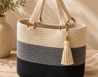 May include: A crochet tote bag featuring a color-blocked design with horizontal stripes in cream, medium gray, and dark charcoal. The bag includes two cream-colored crochet handles attached with wooden rings and a decorative cream tassel accessory.