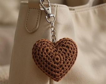 May include: A brown crocheted heart bag charm attached to a cream-colored canvas bag with a silver-toned metal lobster clasp and chain. The text Bag Charm is printed in white at the bottom of the image, which also features a small four-pointed star icon.