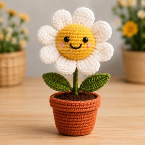 以下が含まれることがあります： テラコッタ色の鉢に入ったかぎ針編みのデイジーの花。白い花びら、笑顔の黄色い中心部、黒い目、ピンクの頬が特徴です。茎には2枚の緑色の葉が付いており、鉢の中には茶色の土が入っています。