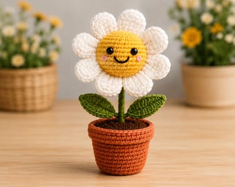 May include: A crocheted daisy flower in a terracotta-coloured pot. The flower has white petals, a yellow centre with a smiling face, black eyes, and pink cheeks. Two green leaves are attached to the stem, and the pot contains brown soil.